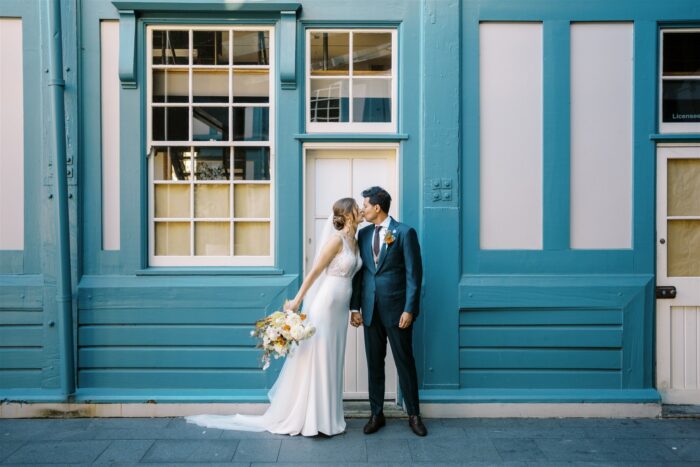 Bride and Groom kissing in front of teal coloured buliding