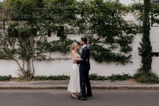 Couple embracing on the street in front of wall covered in foliage.