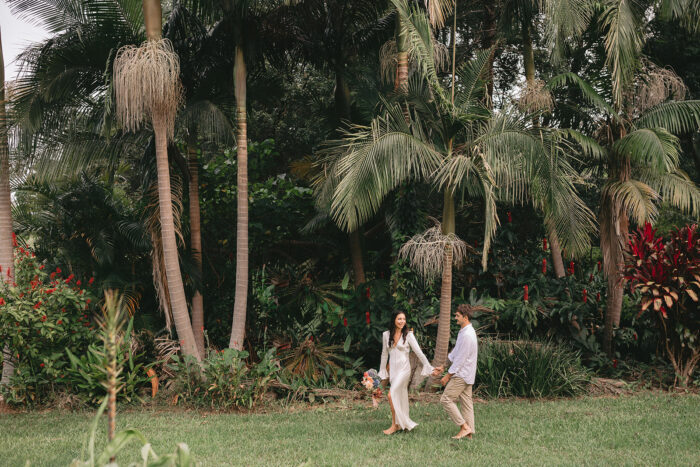Bride holding bouquet and groom hold hands and wander barefoot across lawn with rainforest and palm trees in the background.
