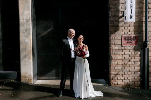 Bride and Groom smiling side by side in front of industrial looking black roller door on suburban street in the sunshine. Bride is holding a bouquet of maroon and red flowers.